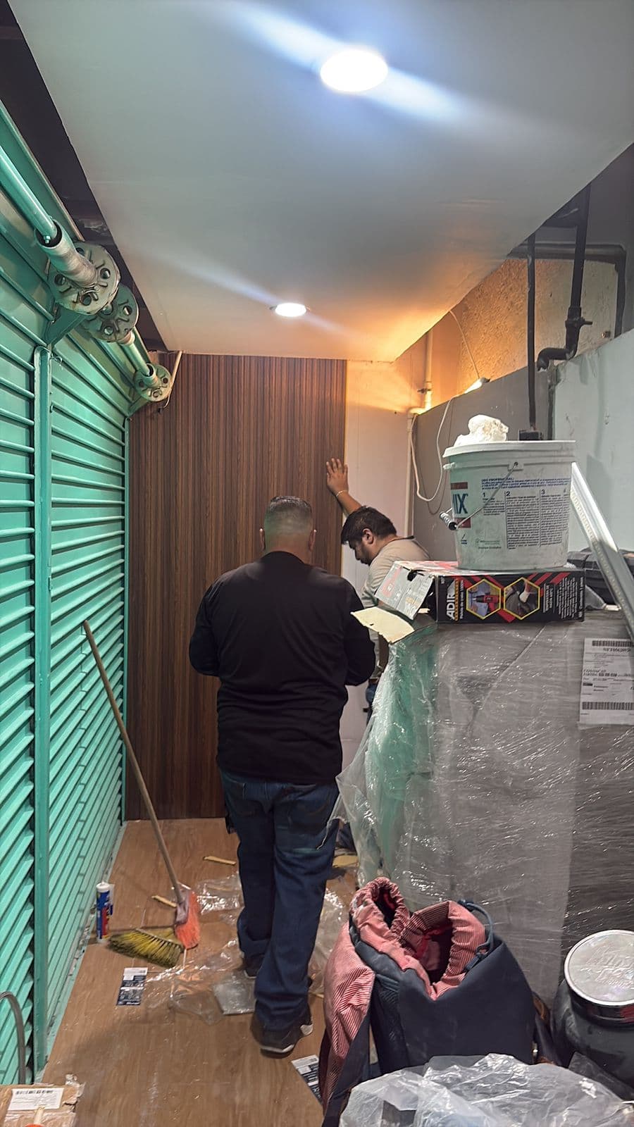 Dos hombres instalan un panel de madera en un pasillo estrecho con materiales de construcción.